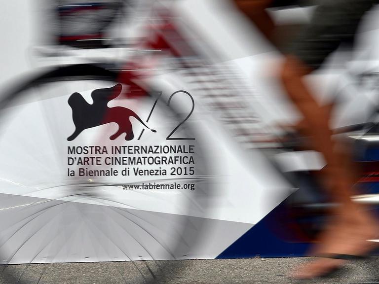 Vor dem offiziellen Logo der 72. Filmfetspiele in Venedig 2015 fährt ein Mann Fahrrad. Schemenhaft sind die Beine und das Fahrrad zu sehen.