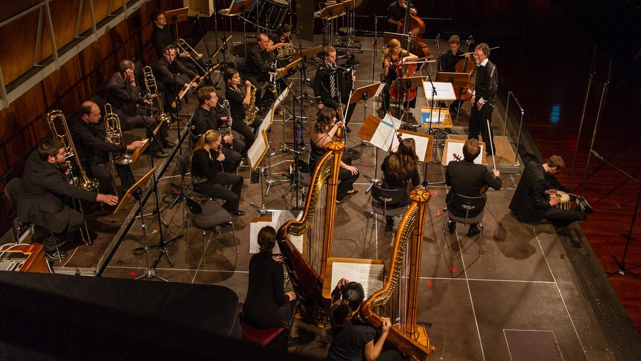 Seitenansicht auf eine Bühne, auf der ein Orchester aus unterschiedlichen Instrumenten sitzt. Vor dem Klangkörper steht ein lächelnder Dirigent, an der Bühnenkante sitzt ein Mann mit einer Trommel. Seitenansicht auf eine Bühne, auf der ein Orchester aus unterschiedlichen Instrumenten sitzt. Vor dem Klangkörper steht ein lächelnder Dirigent, an der Bühnenkante sitzt ein Mann mit einer Trommel.