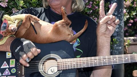 Ein älterer Hippie sitzt mit einer Gitarre und einem kleinen Hund auf dem Arm auf einer Bank und zeigt lachend das Victory-Zeichen