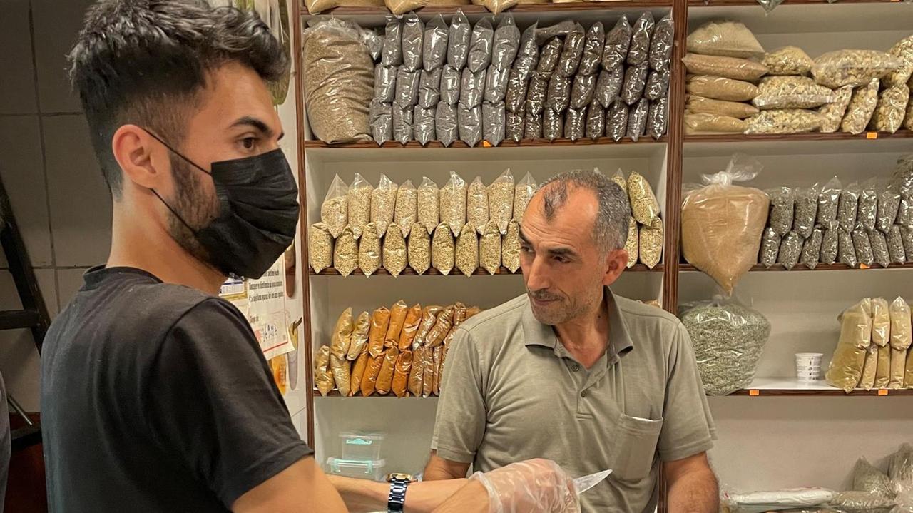 Ein junger Mann in Jeans, schwarzem T-Shirt und Mundschutz steht vor einem Regal mit Gewürzen und einem Verkäufer. Ein junger Mann in Jeans, schwarzem T-Shirt und Mundschutz steht vor einem Regal mit Gewürzen und einem Verkäufer.