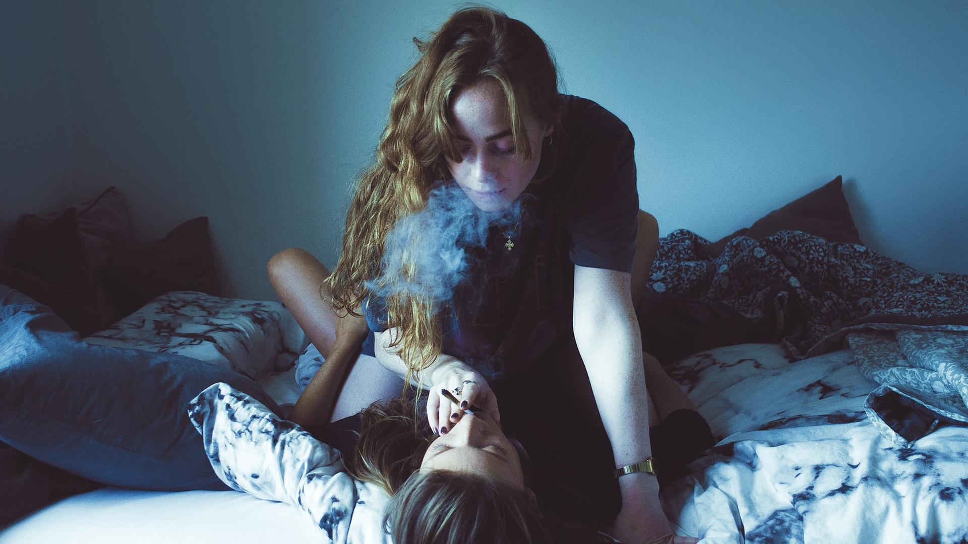 Zwei Frauen auf einem Bett, eine reicht der anderen eine Zigarette an den Mund. Zwei Frauen auf einem Bett, eine reicht der anderen eine Zigarette an den Mund.