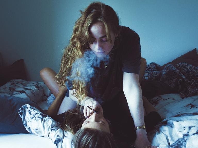 Zwei Frauen auf einem Bett, eine reicht der anderen eine Zigarette an den Mund.