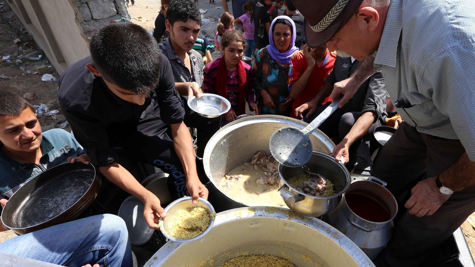 Zahlreiche Menschen stehen um zwei große Töpfe, aus denen Essen ausgegeben wird. Zahlreiche Menschen stehen um zwei große Töpfe, aus denen Essen ausgegeben wird.