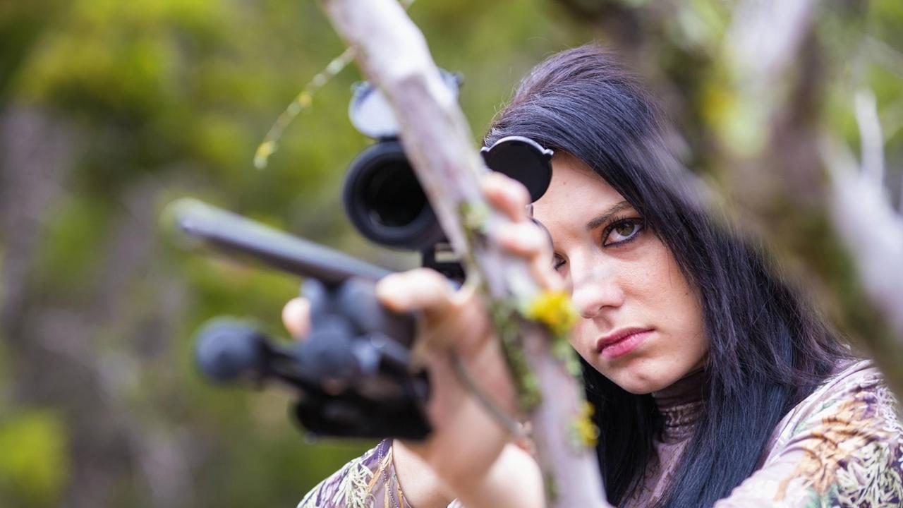 Eine junge Frau mit schwarzen Haaren hält ein Jagdgewehr in der Hand und zielt.