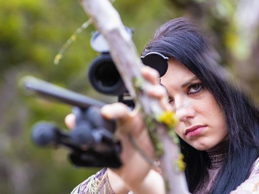 Eine junge Frau mit schwarzen Haaren hält ein Jagdgewehr in der Hand und zielt.