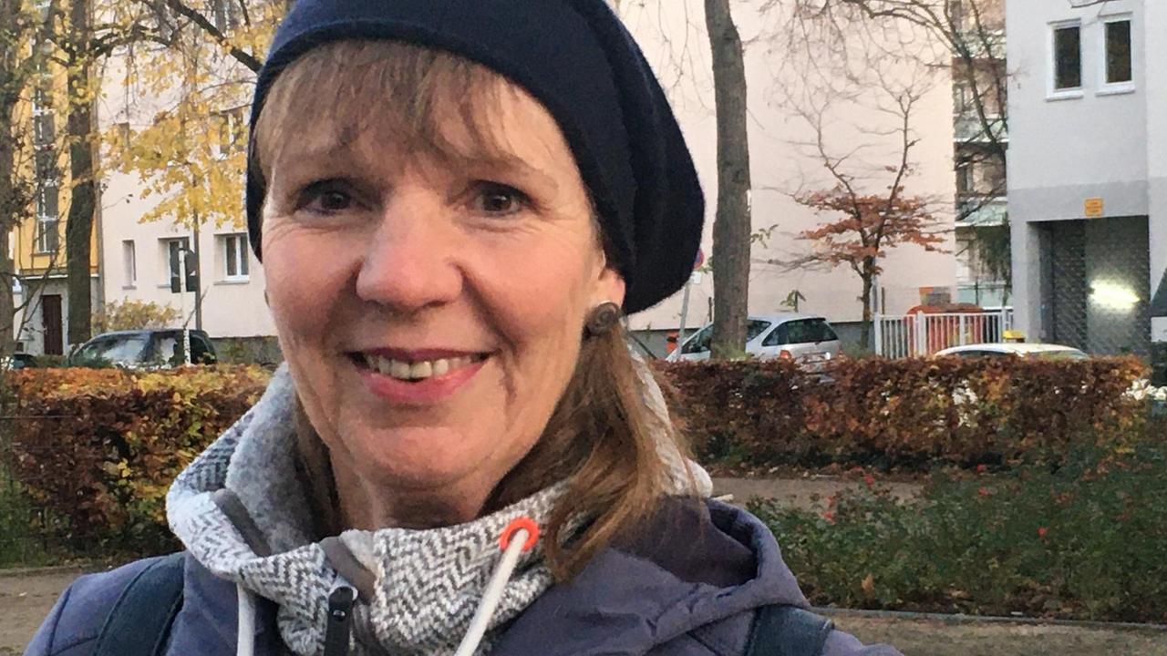 Porträt von Regine Sophia Gartmann, im blauen Anorak mit schwarzer Mütze, im Hintergrund ein kleiner Park mit gelbem Herbstlaub. Porträt von Regine Sophia Gartmann, im blauen Anorak mit schwarzer Mütze, im Hintergrund ein kleiner Park mit gelbem Herbstlaub.