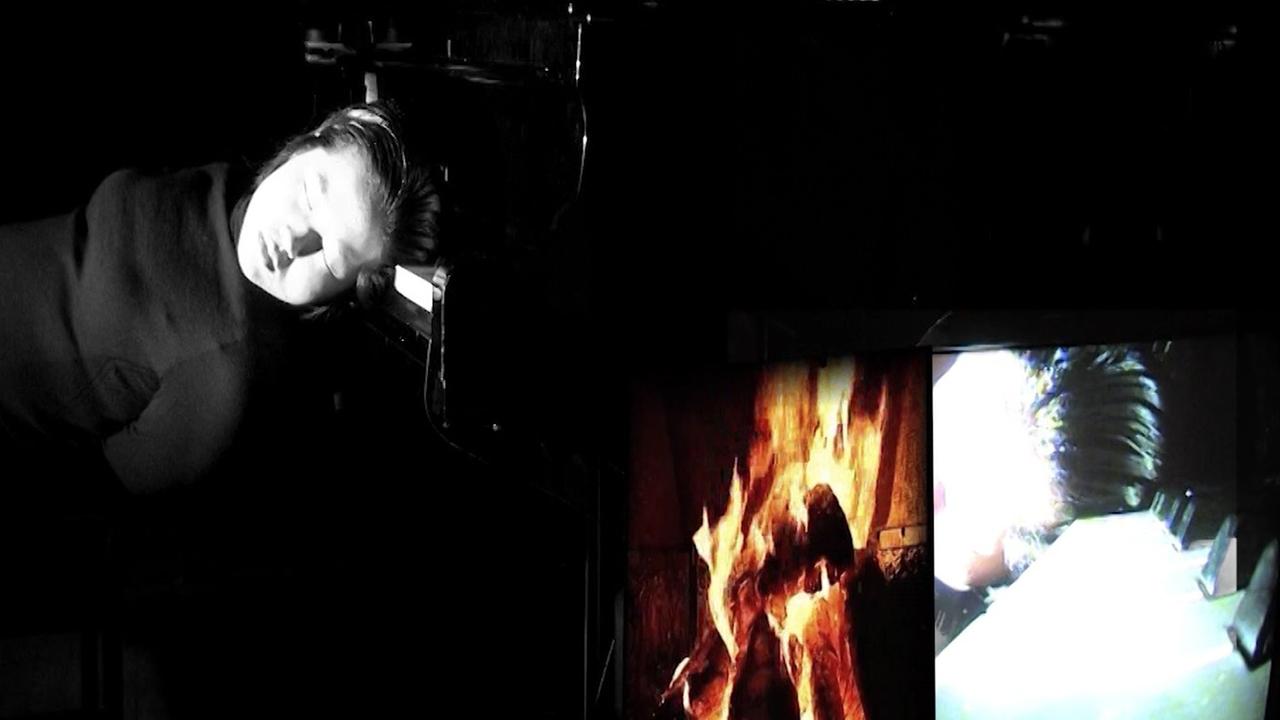 Eine theatralische Szenerie mit dem Kopf der Pianistin, der seitlich auf der Tastatur des Flügels liegt, während auf zwei Monitoren je ein Kaminfeuer und eine Nahaufnahme ihres Kopfes gezeigt werden. Eine theatralische Szenerie mit dem Kopf der Pianistin, der seitlich auf der Tastatur des Flügels liegt, während auf zwei Monitoren je ein Kaminfeuer und eine Nahaufnahme ihres Kopfes gezeigt werden.