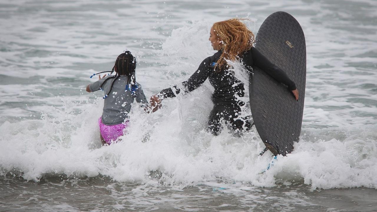 Die Surflehrerin und ihre Schülerin laufen in das spritzende Wasser.