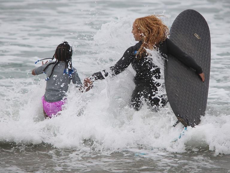 Die Surflehrerin und ihre Schülerin laufen in das spritzende Wasser.