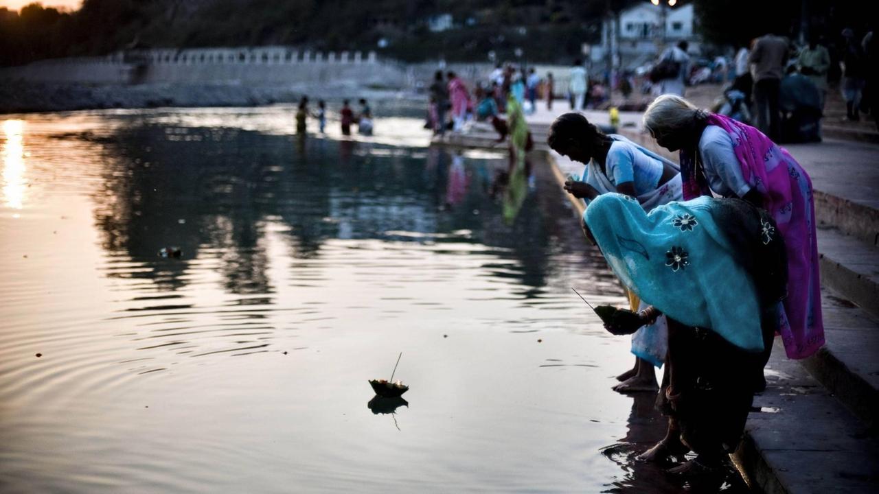 Drei Frauen stehen gebeugt und betend auf Stufen am Rand eines Flusses auf das Wasser legen sie Gaben (Indien, Haridwar)