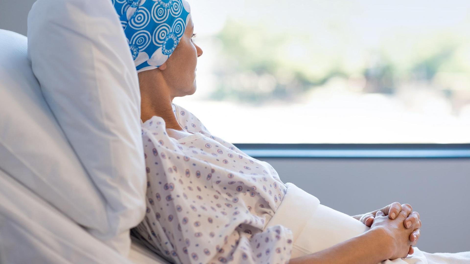 Eine junge Frau mit blauer Kopfbedeckung liegt nachdenklich im Krankenhausbett, hat die Hände ineinander gefaltet und schaut aus dem Fenster raus. Eine junge Frau mit blauer Kopfbedeckung liegt nachdenklich im Krankenhausbett, hat die Hände ineinander gefaltet und schaut aus dem Fenster raus.