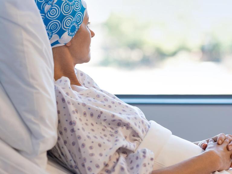 Eine junge Frau mit blauer Kopfbedeckung liegt nachdenklich im Krankenhausbett, hat die Hände ineinander gefaltet und schaut aus dem Fenster raus.