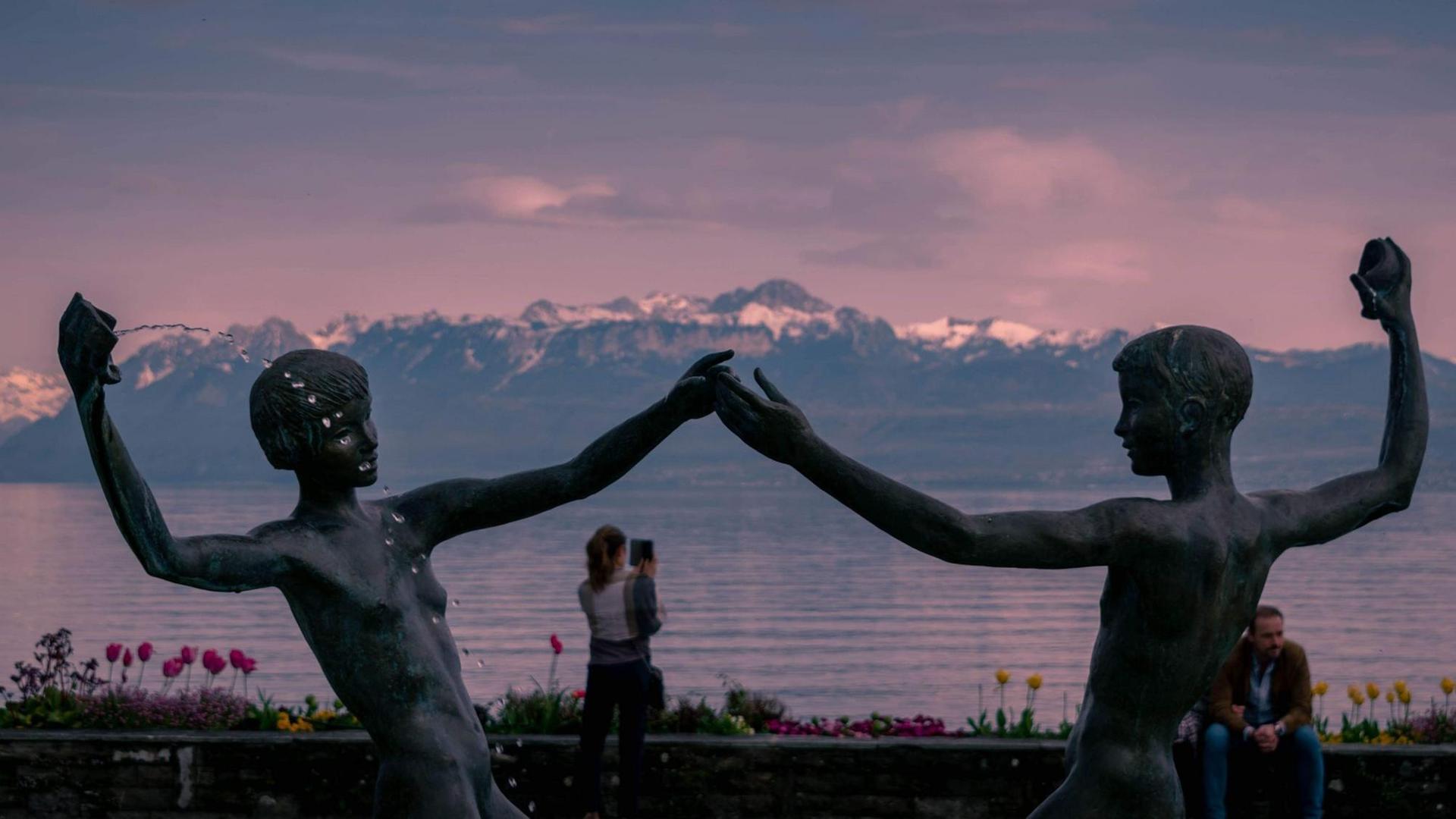 Zwei Brunnenfiguren reichen sich die Hände vor dem rosa Himmel, einem See und einem Alpenpanorama im Hintergrund.
