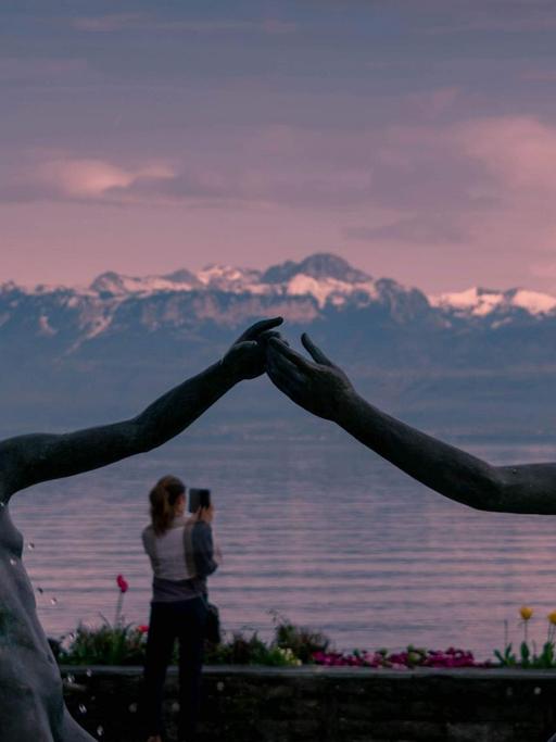 Zwei Brunnenfiguren reichen sich die Hände vor dem rosa Himmel, einem See und einem Alpenpanorama im Hintergrund.