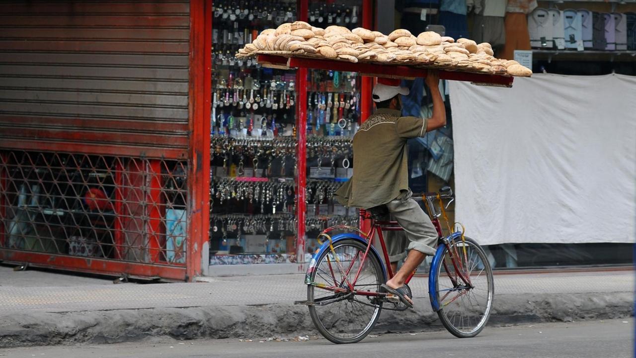 Ein Brotverkäufer balanciert ein großes Brettgestellt voll beladen mit Brot auf seinem Kopf und fährt mit dem Fahrrad durch eine Straße in Kairo zur Kundschaft. Ein Brotverkäufer balanciert ein großes Brettgestellt voll beladen mit Brot auf seinem Kopf und fährt mit dem Fahrrad durch eine Straße in Kairo zur Kundschaft.