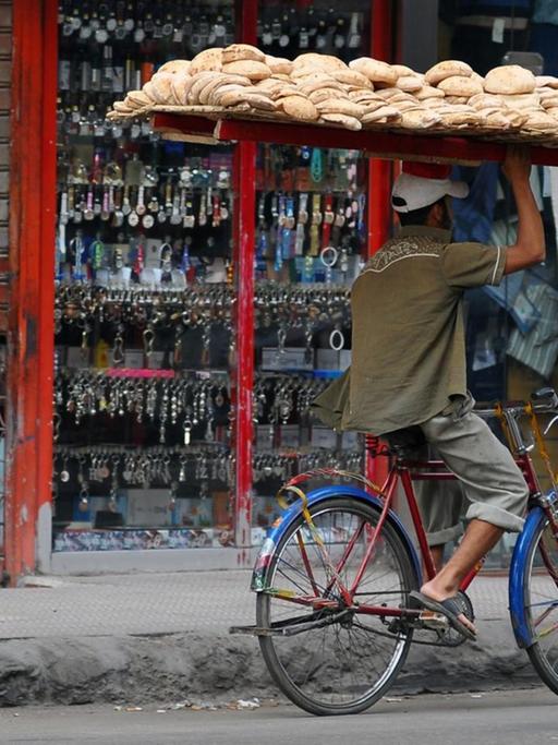 Ein Brotverkäufer balanciert ein großes Brettgestellt voll beladen mit Brot auf seinem Kopf und fährt mit dem Fahrrad durch eine Straße in Kairo zur Kundschaft.