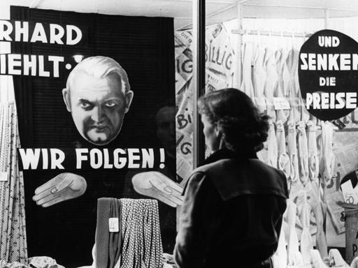 Eine Frau steht vor einem Schaufenster. In dem Schaufenster steht "Erhard befiehlt - wir folgen! und senken die Preise". Das Foto ist von circa 1949/50.