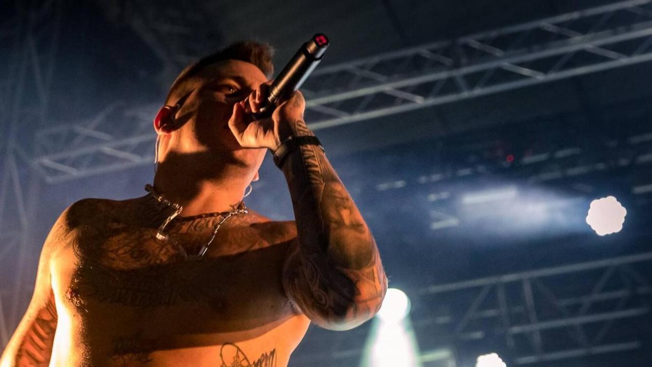 Der Rapper Gzuz steht auf einer Bühne und singt in ein Mikrofon, er wird von hintern von Scheinwerfern angeleutet. Der Rapper Gzuz steht auf einer Bühne und singt in ein Mikrofon, er wird von hintern von Scheinwerfern angeleutet.