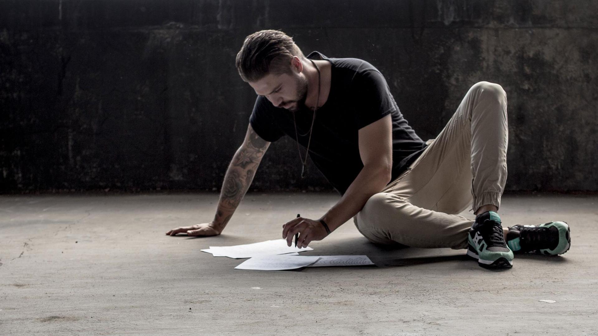Ein Mann sitzt auf einem Betonboden und schreibt auf einigen Blättern, die er vor sich ausgebreitet hat.