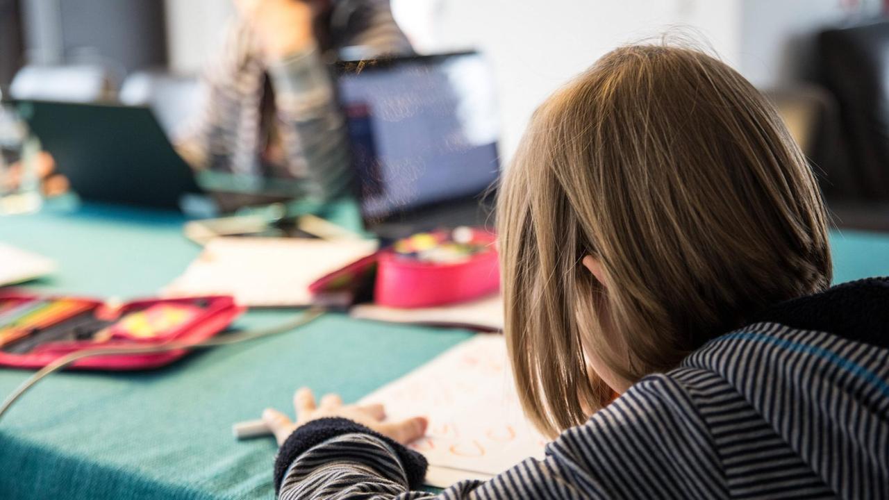 Ein Schulkind sitzt an einem Tisch und macht Hausaufgaben im Homeschooling, im Hintergrund sitzt die Mutter des Kindes an einem Laptop und macht Homeoffice. Auf dem Tisch liegen Schulutensilien und ein weiterer aufgeklappter Laptop. Ein Schulkind sitzt an einem Tisch und macht Hausaufgaben im Homeschooling, im Hintergrund sitzt die Mutter des Kindes an einem Laptop und macht Homeoffice. Auf dem Tisch liegen Schulutensilien und ein weiterer aufgeklappter Laptop.