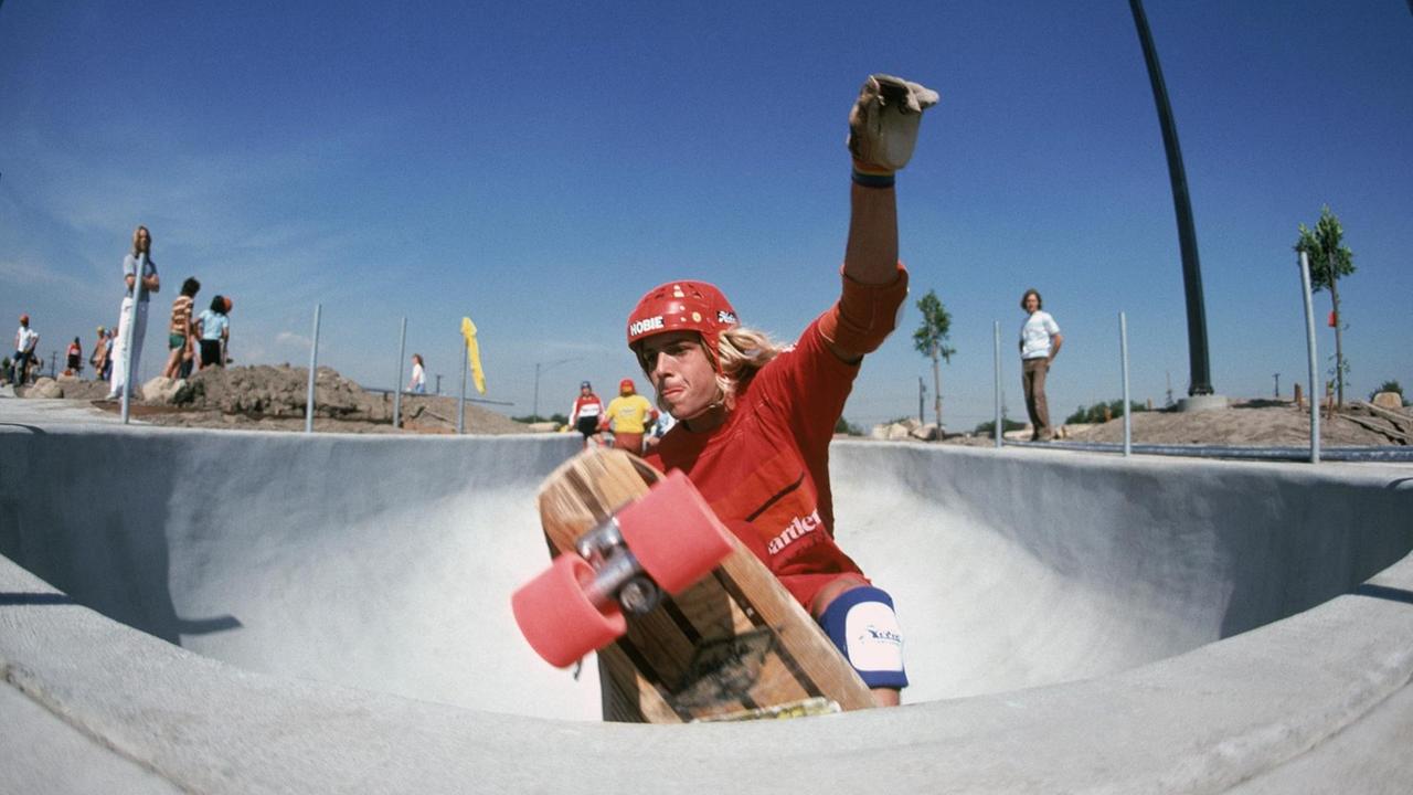 Ein Foto aus den 70ern mit einem großen Weitwinkel zeigt einen Skateboarder der gerade an der abgerundeten Oberkante eines Beton-Pools. Im Hintergrund sind Palmen zu sehen.