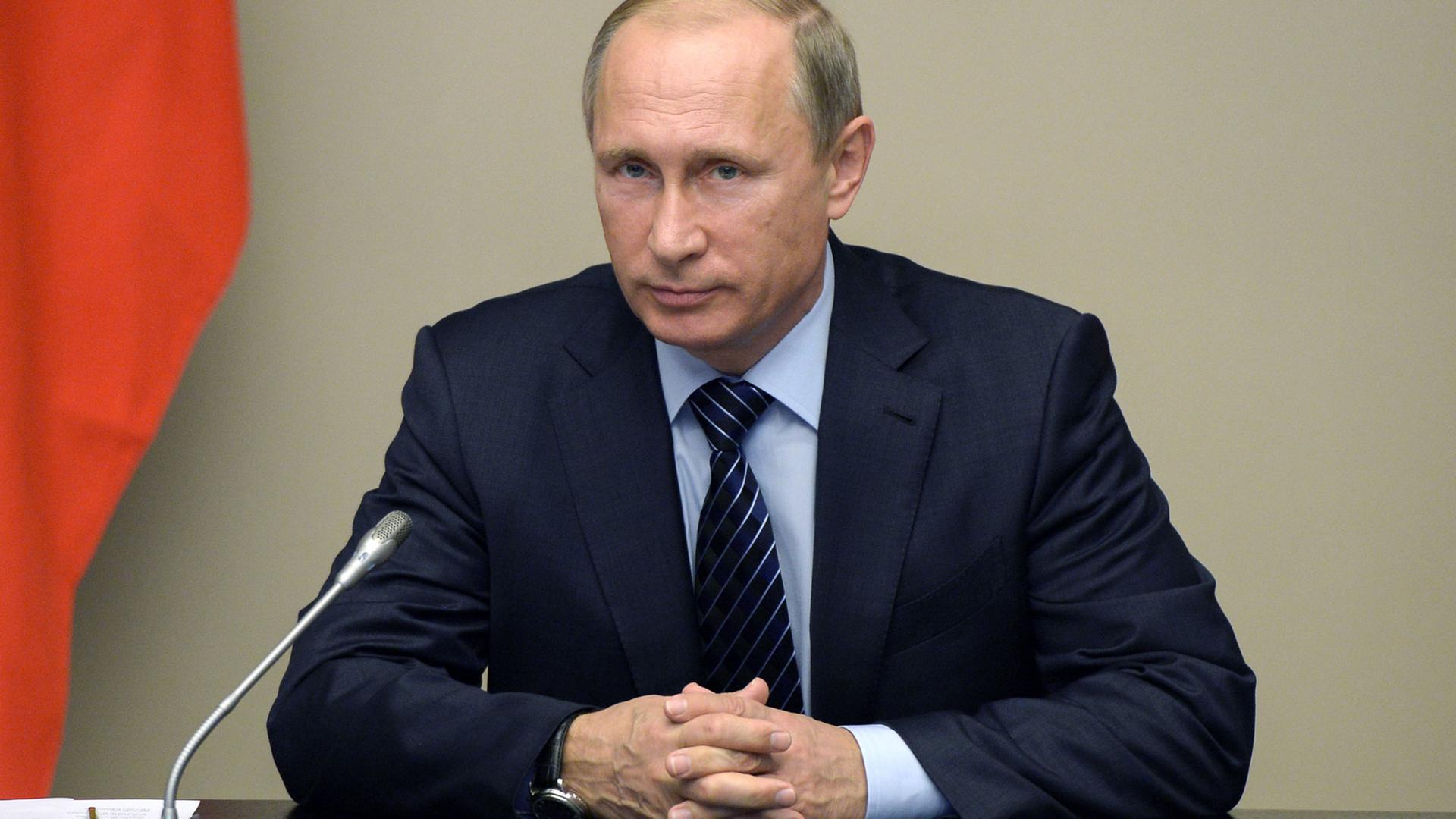 Der russische Präsident Wladimir Putin sitzt am 25.09.2015 in seinem Amtssitz in Nowo-Ogarjowo bei einem Treffen des Sicherheitsrats der Russischen Föderation an einem Tisch, auf dem sich ein Mikrofon und Schreitutensilien befinden; im Hintergrund ein rotes Stück Fahne. Der russische Präsident Wladimir Putin sitzt am 25.09.2015 in seinem Amtssitz in Nowo-Ogarjowo bei einem Treffen des Sicherheitsrats der Russischen Föderation an einem Tisch, auf dem sich ein Mikrofon und Schreitutensilien befinden; im Hintergrund ein rotes Stück Fahne.