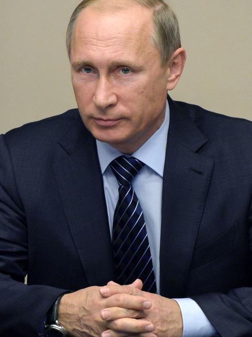 Der russische Präsident Wladimir Putin sitzt am 25.09.2015 in seinem Amtssitz in Nowo-Ogarjowo bei einem Treffen des Sicherheitsrats der Russischen Föderation an einem Tisch, auf dem sich ein Mikrofon und Schreitutensilien befinden; im Hintergrund ein rotes Stück Fahne.