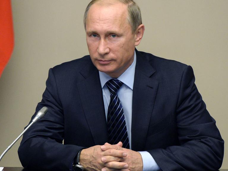 Der russische Präsident Wladimir Putin sitzt am 25.09.2015 in seinem Amtssitz in Nowo-Ogarjowo bei einem Treffen des Sicherheitsrats der Russischen Föderation an einem Tisch, auf dem sich ein Mikrofon und Schreitutensilien befinden; im Hintergrund ein rotes Stück Fahne.