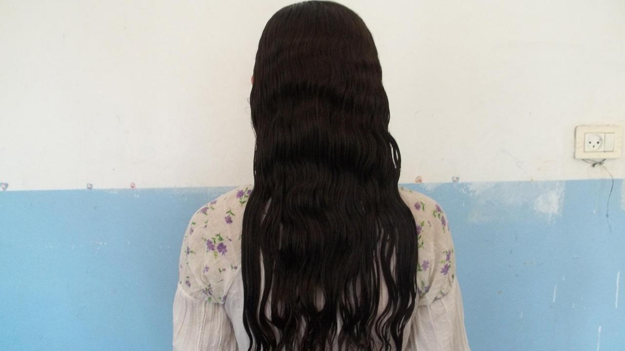 Eine Frau mit langen schwarzen Haaren steht mit dem Gesicht zur Wand. Zu sehen ist ihr Rücken.