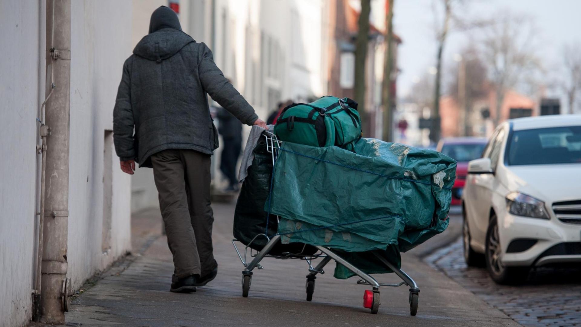 Ein Mann schiebt einen Einkaufswagen, samt Planen, Rucksack und Tasche durch die Stadt Ein Mann schiebt einen Einkaufswagen, samt Planen, Rucksack und Tasche durch die Stadt