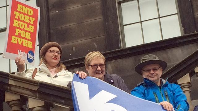 Auf einer Außentreppe stehen drei Unabhängigkeits-Befürworter mit Yes-Plakat und einem Banner mit Schotten-Karo.