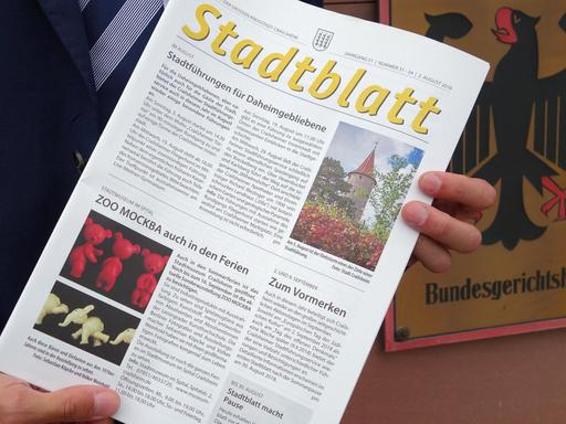 Eine Ausgabe des Amtsblattes der Stadt Crailsheim wird von zwei Händen gehalten. Dahinter das Schild des Bundesgerichtshofes.