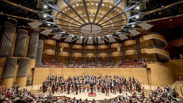 Ein Orchester in großer Besetzung nimmt stehend Beifall in der Kölner Philharmonie an.