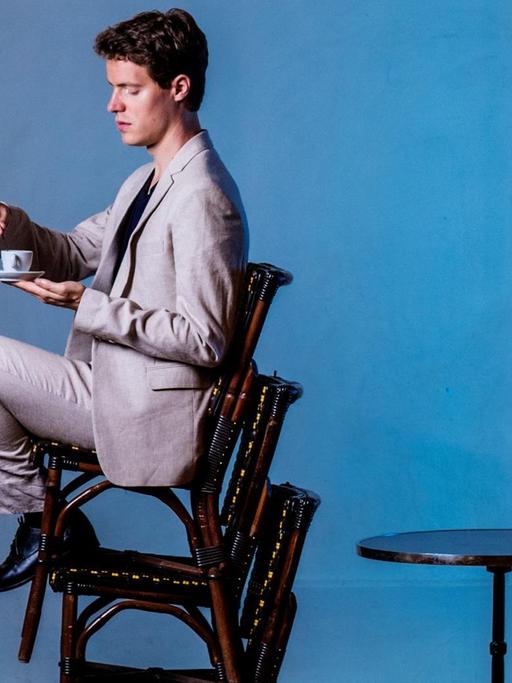 Ein Mann sitzt auf dem obersten von drei übereinander gestapelten Stühlen und hält eine Teetasse vor sich. Man sieht ihn seitlich, der Hintergrund ist bläulich, er trägt einen hellen Anzug.
