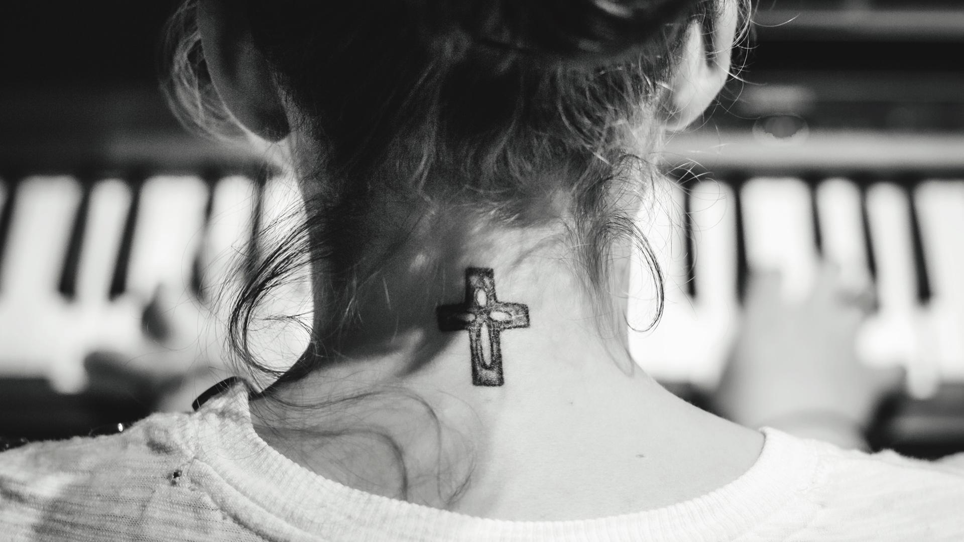 Eine junge Frau mit hochgesteckten Haaren sitzt am Klavier und spielt: Gezeigt wird sie in der Rückenansicht, so dass an ihrem Hals das Tattoo eines christlichen Kreuzes sichtbar wird.