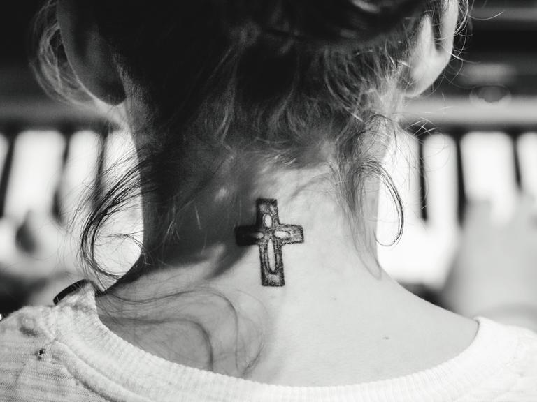 Eine junge Frau mit hochgesteckten Haaren sitzt am Klavier und spielt: Gezeigt wird sie in der Rückenansicht, so dass an ihrem Hals das Tattoo eines christlichen Kreuzes sichtbar wird.