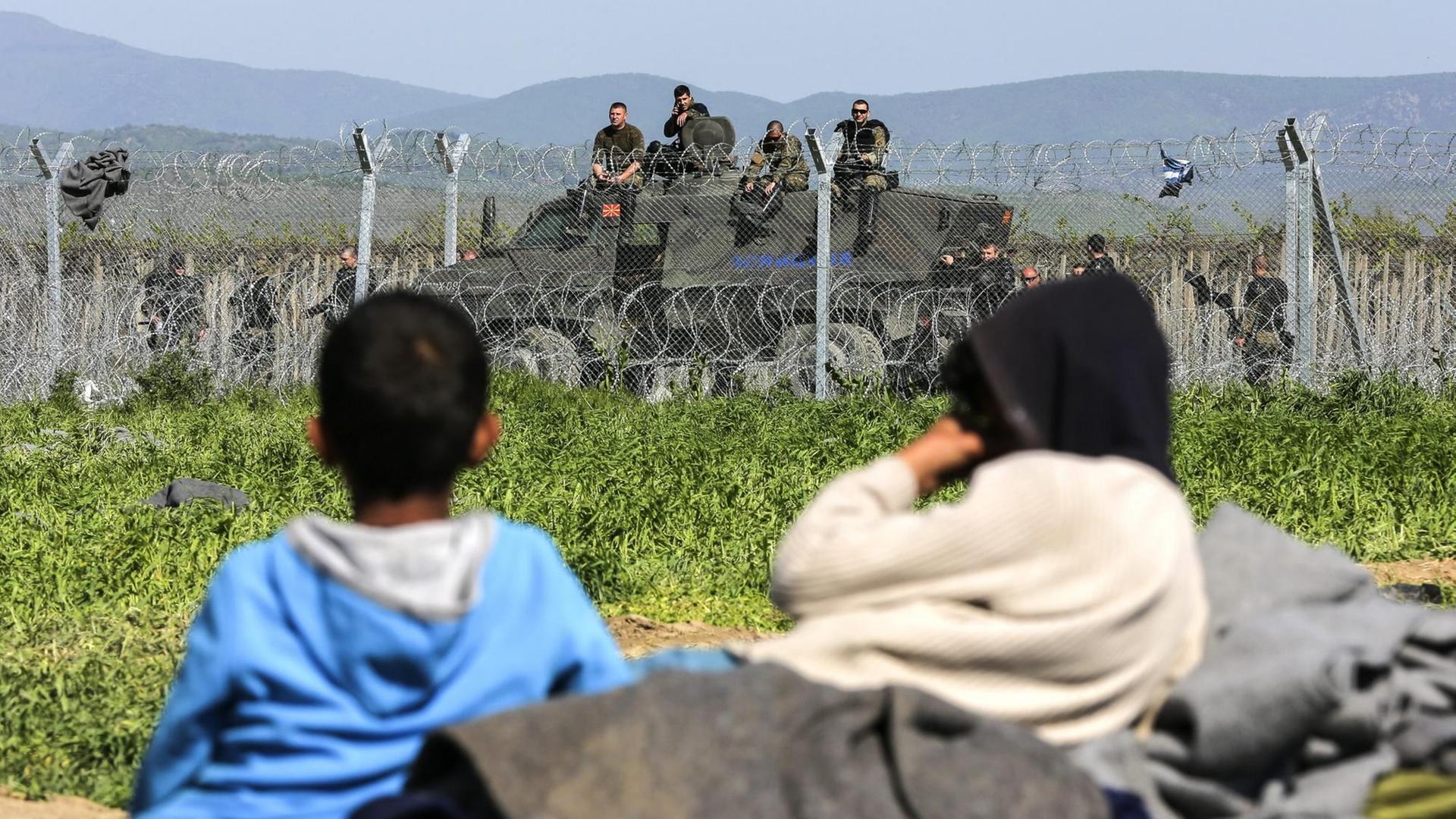 Zwei Flüchtlingskinder liegen im Gras vor einem Stacheldrahtzaun, der Griechenland von Mazedonien trennt. Im Hintergrund ist ein Panzer mit Grenzschutzbeamten zu sehen. Zwei Flüchtlingskinder liegen im Gras vor einem Stacheldrahtzaun, der Griechenland von Mazedonien trennt. Im Hintergrund ist ein Panzer mit Grenzschutzbeamten zu sehen.
