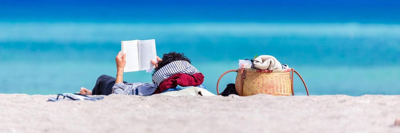 Das Bild zeigt einen Strand. Im Hintergrund ist eine im Sand liegende Frau zu sehen, die ein Buch in der Hand hält und liest. Dahinter ist intensiv blau Himmel und Meer zu sehen. Das Bild zeigt einen Strand. Im Hintergrund ist eine im Sand liegende Frau zu sehen, die ein Buch in der Hand hält und liest. Dahinter ist intensiv blau Himmel und Meer zu sehen.