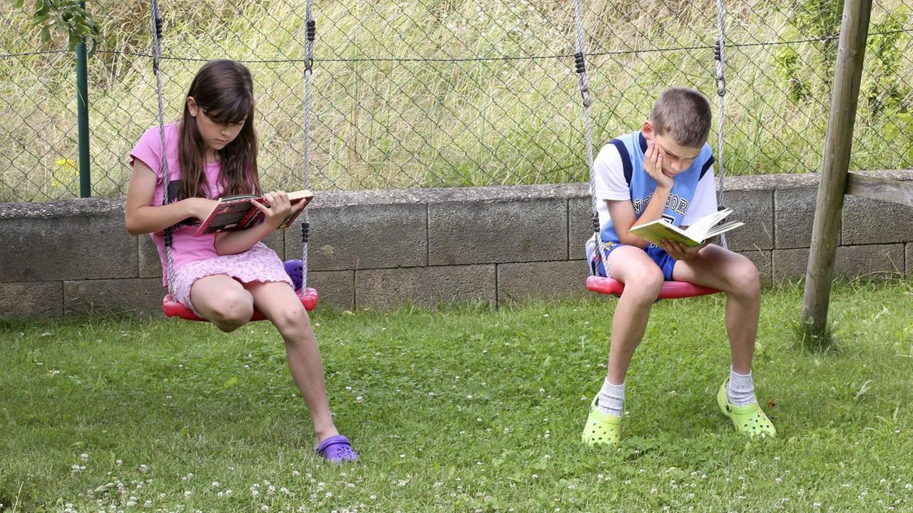 Ein Mädchen und ein Junge lesen Bücher und sitzen dabei auf Schaukeln Ein Mädchen und ein Junge lesen Bücher und sitzen dabei auf Schaukeln