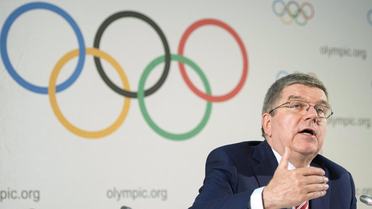 Bach spricht in ein Mikrofon und gestikuliert. An der Wand im Hintergrund sieht man die Olympischen Ringe. Bach spricht in ein Mikrofon und gestikuliert. An der Wand im Hintergrund sieht man die Olympischen Ringe.