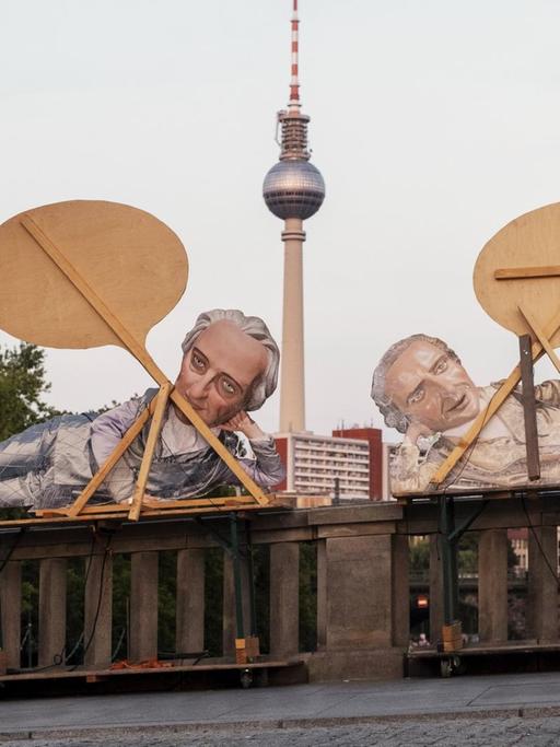 Werbung für ein Berliner Theater mittels Pappfiguren Goethe und Schiller an der Monbijoubrücke. Im Vordergrund ist ein junges Paar zu sehen. Dahinter der Fernsehturm. 