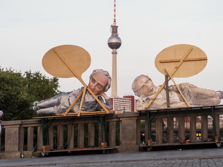 Werbung für ein Berliner Theater mittels Pappfiguren Goethe und Schiller an der Monbijoubrücke. Im Vordergrund ist ein junges Paar zu sehen. Dahinter der Fernsehturm. 