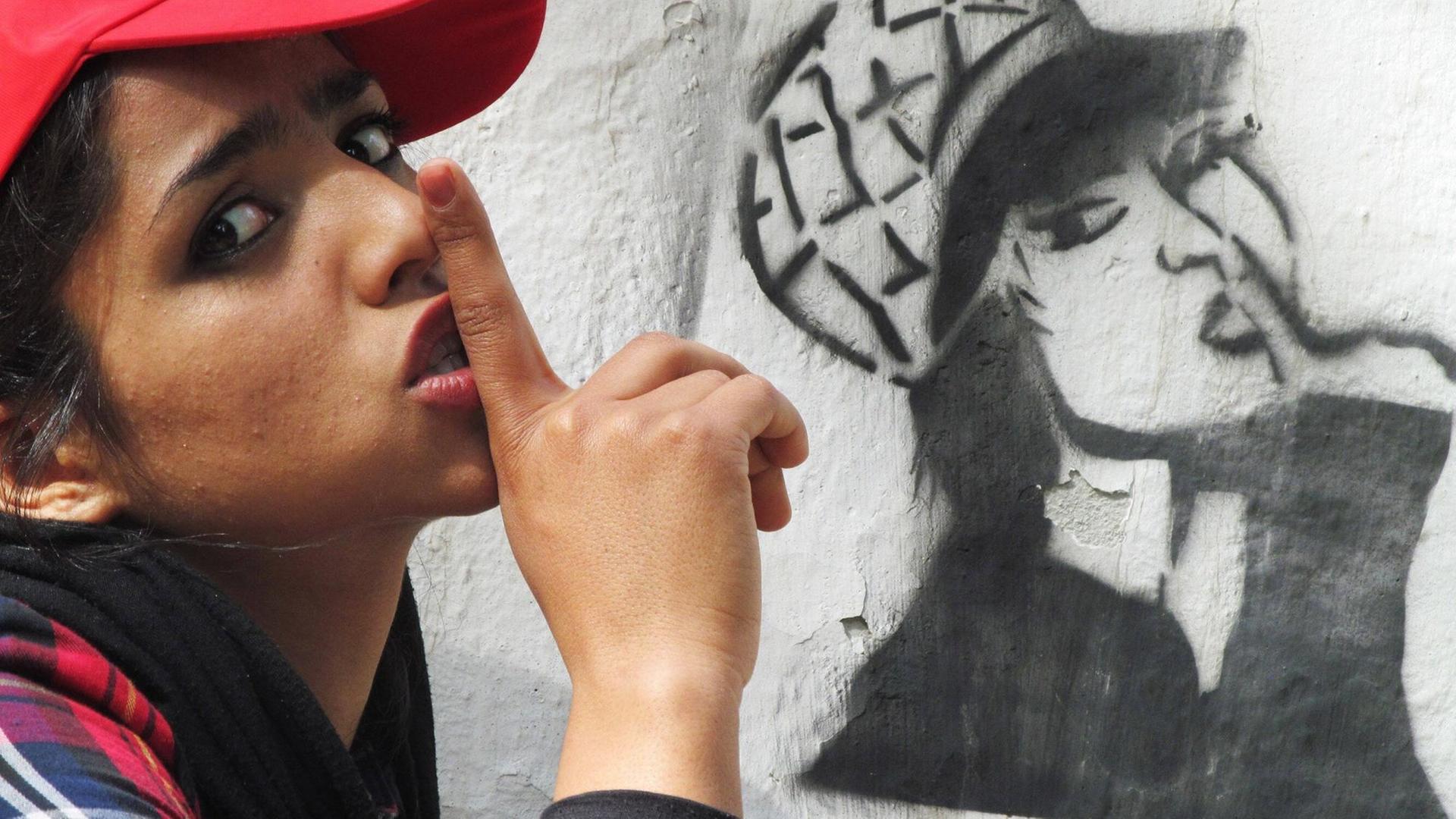 Sonita Alizadeh steht im Profil vor einer weißen Wand, auf der ein Graffito gesprüht ist. Sie trägt eine rote Basecap und hält den Zeigefinger ihrer rechten Hand an den Mund. Das Graffito zeigt die gleiche Pose. Sonita Alizadeh steht im Profil vor einer weißen Wand, auf der ein Graffito gesprüht ist. Sie trägt eine rote Basecap und hält den Zeigefinger ihrer rechten Hand an den Mund. Das Graffito zeigt die gleiche Pose.