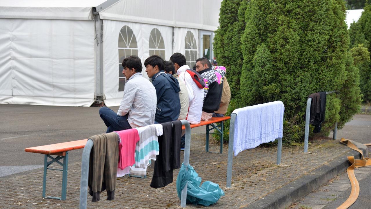 Bewohner der Außenstelle der Aufnahmeeinrichtung für Asylsuchende (AfA) Ingelheim auf dem Flughafen Frankfurt-Hahn in Lautzenhausen (Rhein-Hunsrück-Kreis) sitzen am 17.08.2015 auf einer Bank vor einem Zelt.