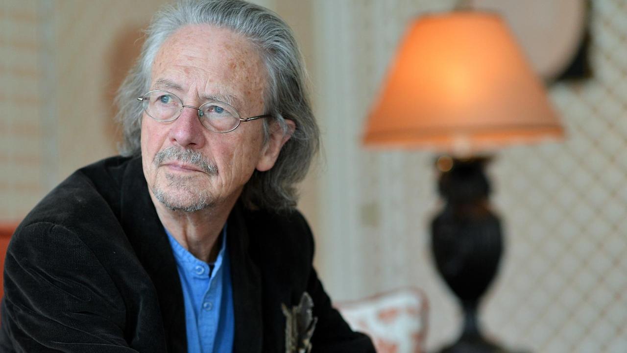Peter Handke sitzt auf einem roten Sofa, hinter ihm steht eine Tischlampe. Peter Handke sitzt auf einem roten Sofa, hinter ihm steht eine Tischlampe.
