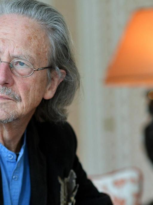 Peter Handke sitzt auf einem roten Sofa, hinter ihm steht eine Tischlampe.