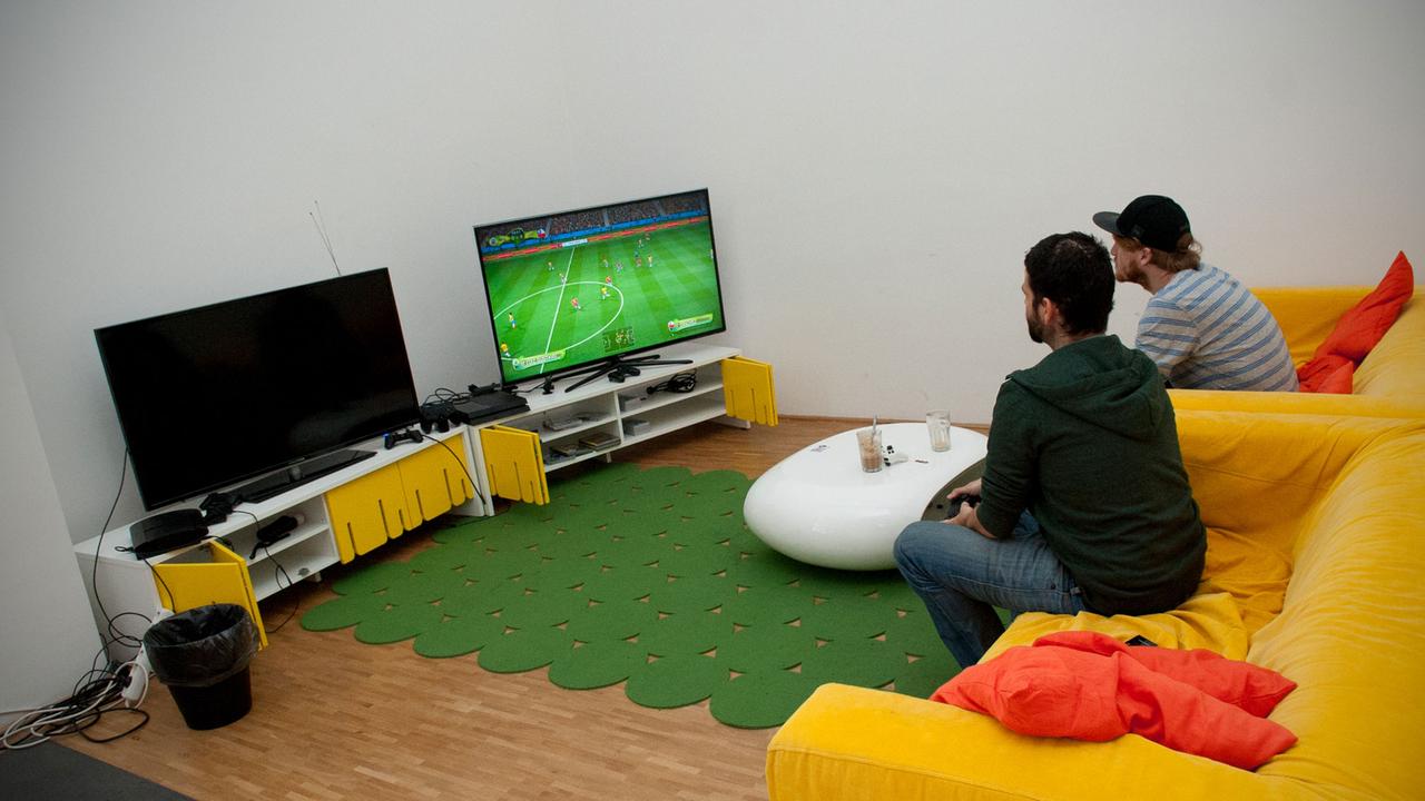 Zwei Angestellte sitzen auf einem gelben Sofa in der Computerspiele-Firma Wooga in Berlin und spielen im Spieleraum mit einer Playstation.