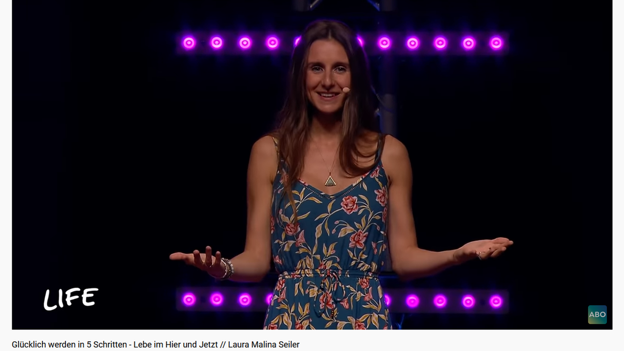 Ein Screenshot aus dem YouTube Video "Glücklich werden in 5 Schritten - Lebe im Hier und Jetzt // Laura Malina Seiler": Die junge Frau steht mit ausgebreiteten Armen auf einer großen Bühne und spricht zu ihrem Publikum Ein Screenshot aus dem YouTube Video "Glücklich werden in 5 Schritten - Lebe im Hier und Jetzt // Laura Malina Seiler": Die junge Frau steht mit ausgebreiteten Armen auf einer großen Bühne und spricht zu ihrem Publikum