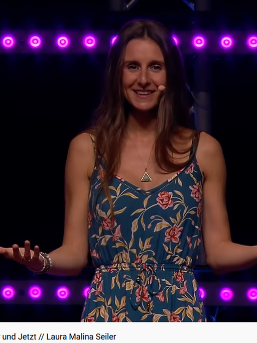 Ein Screenshot aus dem YouTube Video "Glücklich werden in 5 Schritten - Lebe im Hier und Jetzt // Laura Malina Seiler": Die junge Frau steht mit ausgebreiteten Armen auf einer großen Bühne und spricht zu ihrem Publikum