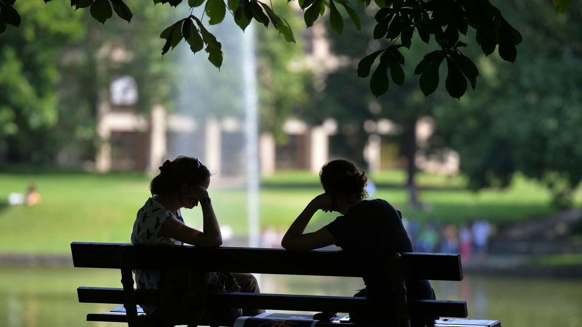Zwei Frauen sitzen im Schatten auf einer Parkbank, nur ihre Silhouetten sind zu erkennen. Zwei Frauen sitzen im Schatten auf einer Parkbank, nur ihre Silhouetten sind zu erkennen.
